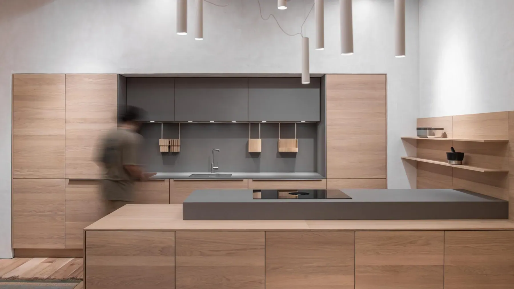 A warm minimal kitchen in the Espai Rö showroom, with a stainless steel island, oak cabinetry and a glazed wall onto a courtyard.